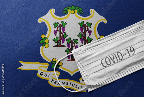 Face Medical Surgical White Mask with COVID-19 inscription lying on Connecticut State Flag. Coronavirus in Connecticut  USA