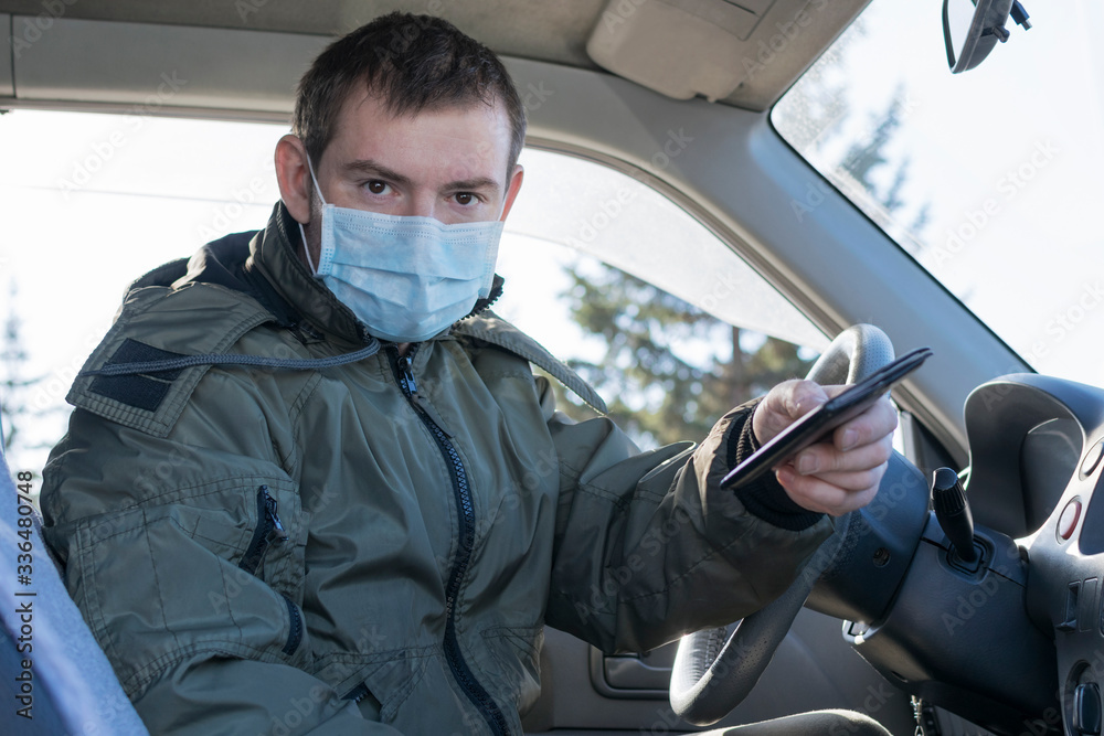 A young man is sitting in a car wearing a medical mask and holding out an identity card for verification during quarantine, epedemia. A young man is sitting in a car wearing a medical mask and holding out an identity card for verification during quarantine, epedemia.