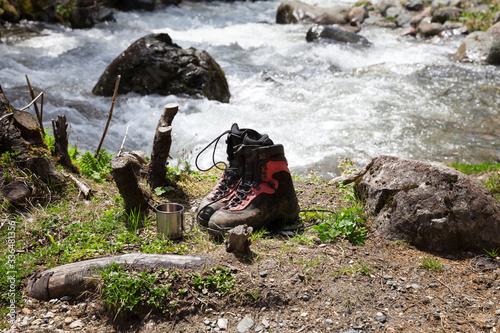 Pair of dirty hiking boots and metal touristic mug on glade with grass near mountain stream