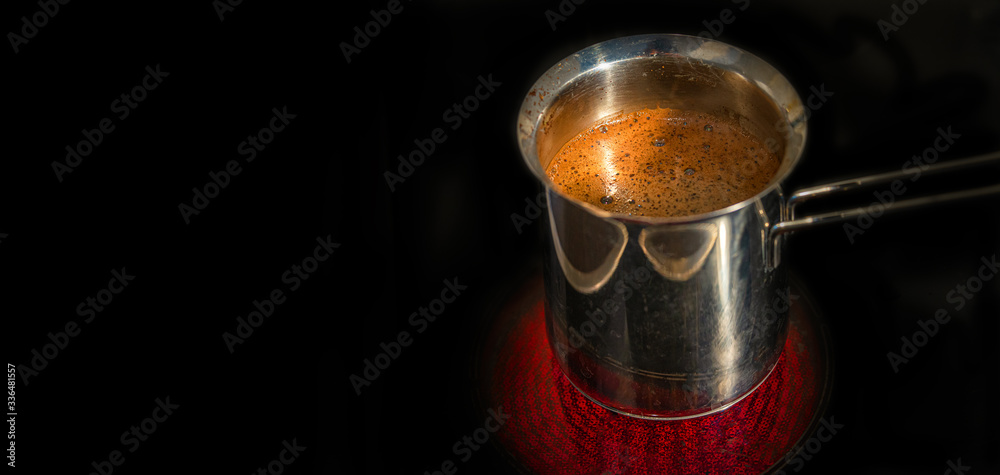 Banner with stainless steel cezve with coffee in process of making standing at hot and red illuminated electrical stove in darkness with copy space, closeup, details