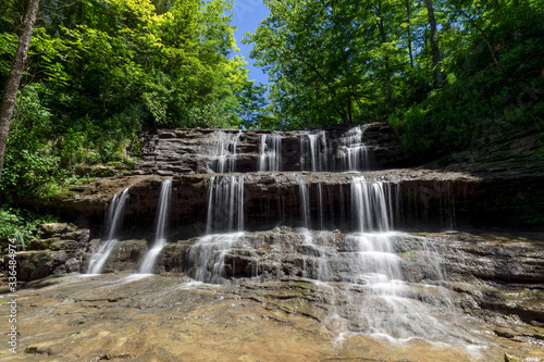 Rivulets of falling water over rock ledges are topped by emerald green leaves and a deep blue sky on a summer day at Fallsville Falls  a beautiful tiered waterfall near Hillsboro  Ohio.