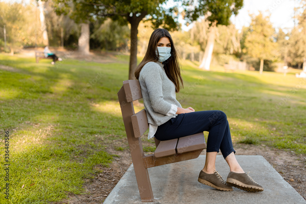 Young woman outdoor taking her medical face mask off. Social distancing sitting on a bench, isolated from other people. Young woman outdoor taking her medical face mask off. Social distancing sitting on a bench, isolated from other people.