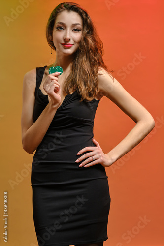 Brunette girl in black dress. She is holding two green chips, smiling, hand on hip, posing on colorful background. Poker, casino. Close-up