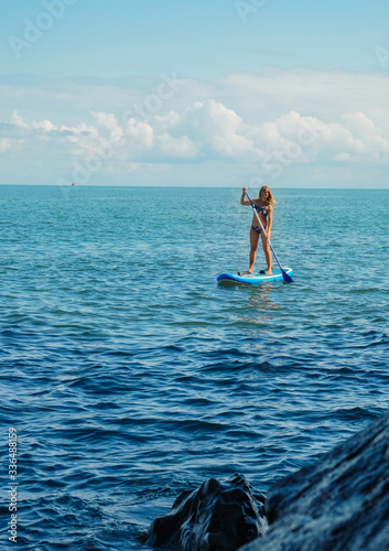 SUP Stand up paddle board. Blond girl stand with paddle in the sea near stone beach. Blue sea with waves Mtsvane Kontskhi Beach, Batumi, Georgia.