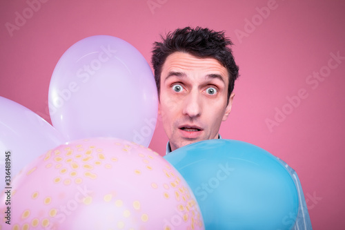 Amazed confused young man look straight and pose with colorful balloons in front. Isolated over pink background. © Anton