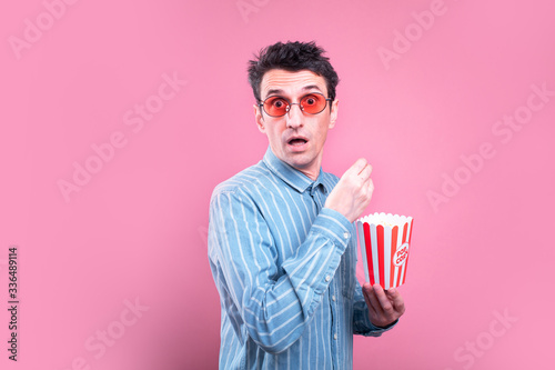 Side view of amazed young man hold small popcorn bucket and look on camera through sunglasses. Eating snacks during movie. Isolated over pink background.