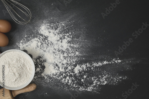 Wooden dough roller, a cup of flour and whisk on the black table with flour spread.