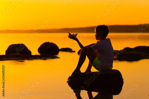 Little boy with paper ship in hands. Beautiful orange sunset. Origami. River. Lake.