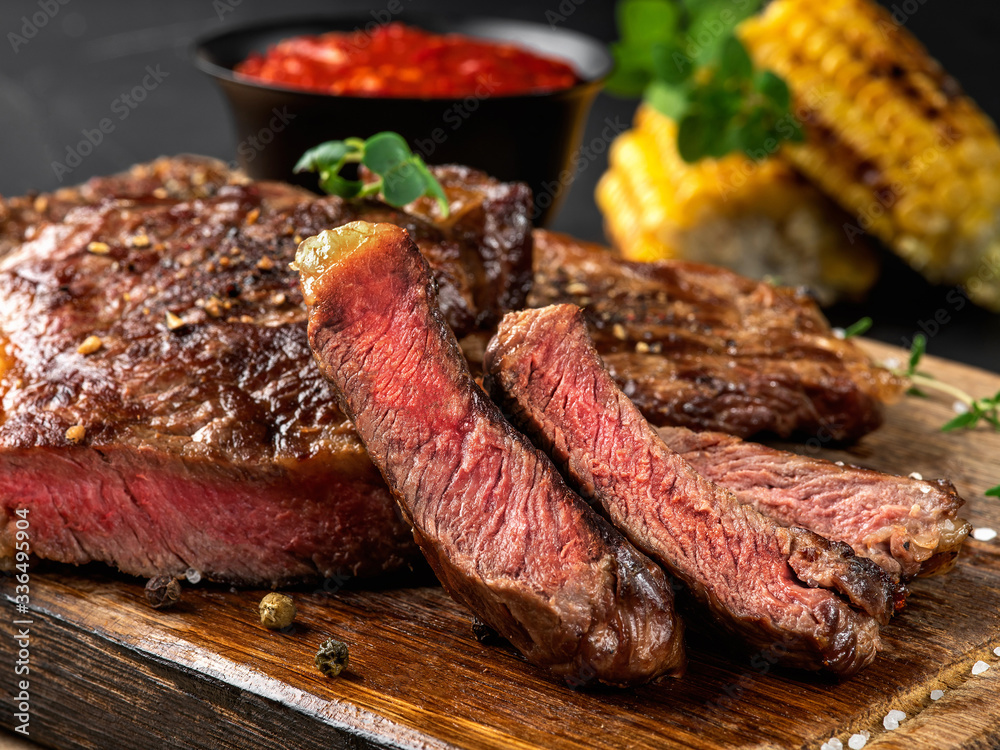 Sliced, fried, spiced steaks with herbs on wooden board, grilled corn, red sauce in small dark bowl on a black background. Close-up shot. Side view.