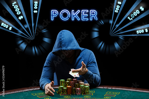 Man in blue hoodie showing aces, sitting at playing table with stacks of chips. Black background with neon dollar amounts, inscription poker. Casino photo