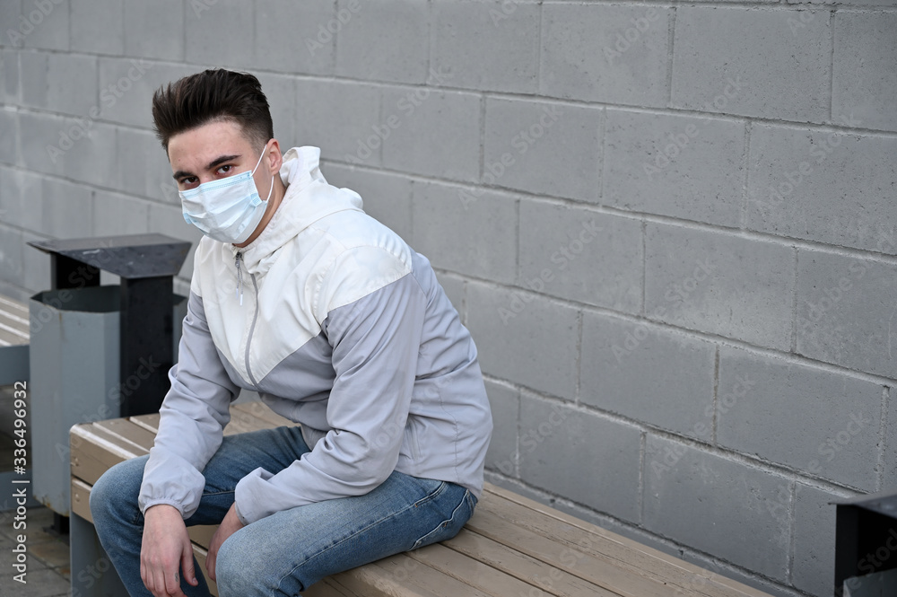 Photo of a Caucasian young brunette man in a medical protective mask in a light jacket outdoors against a gray wall in the city on a sunny day Photo of a Caucasian young brunette man in a medical protective mask in a light jacket outdoors against a gray wall in the city on a sunny day