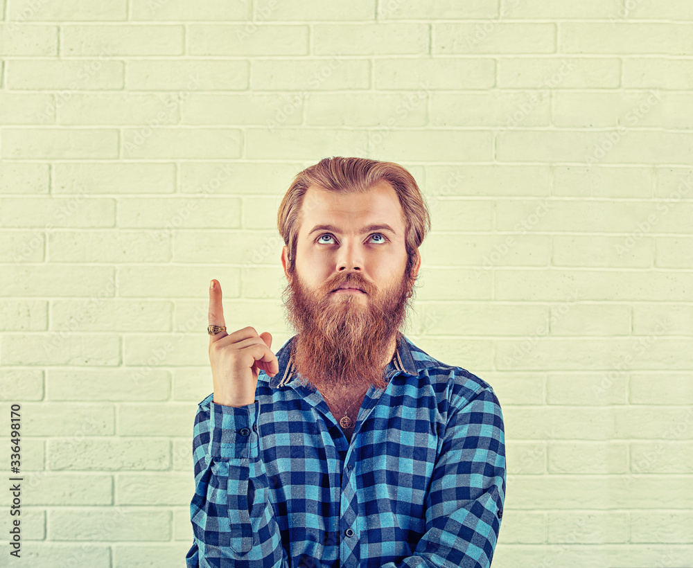 Portrait of a thoughtful wearing casual blue shirt man pointing finger up isolated on a white brick wall background. Yellow Toned image, horizontal. Neutral human emotion, face expression Portrait of a thoughtful wearing casual blue shirt man pointing finger up isolated on a white brick wall background. Yellow Toned image, horizontal. Neutral human emotion, face expression