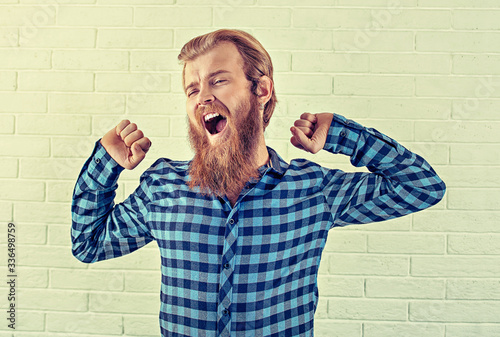 Tired. Portrait of a bearded guy yawning stretching hands open mouth gesture isolated white brick wall background. Yellow Toned image, horizontal. Positive human emotion, face expression