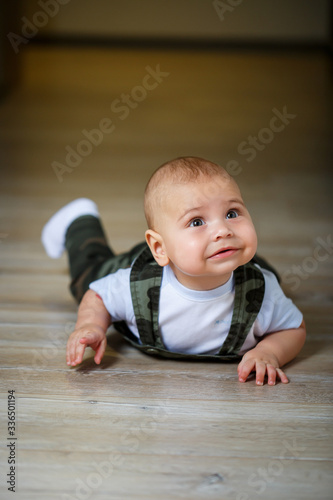 little boy aged 8 months in overalls, a white shirt and white socks crawling on the floor and smiling