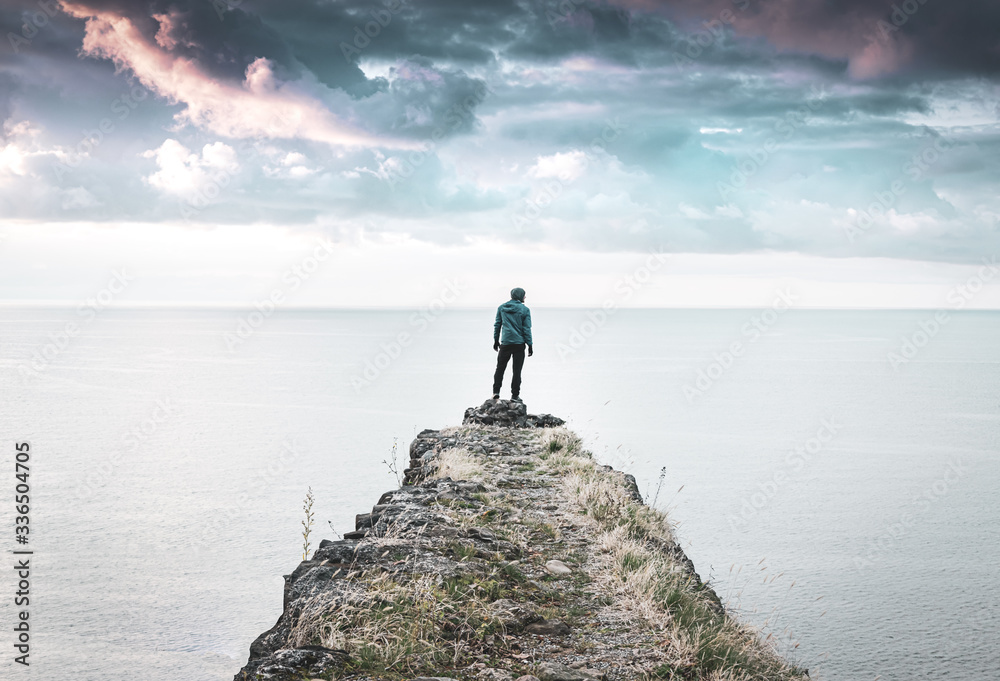 Person stands alone and looks around on the edge of  the cliff with vast sea and dramatic sky in the background.  Concept of solitude, adventuree and lifestyle. 2020