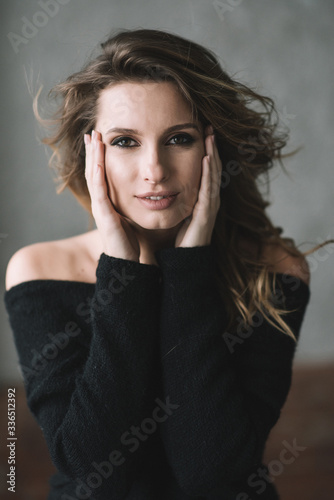 A beautiful woman with curls and full lips in a black sweater holds her face with her hands and looks at the camera. Soft selective focus.