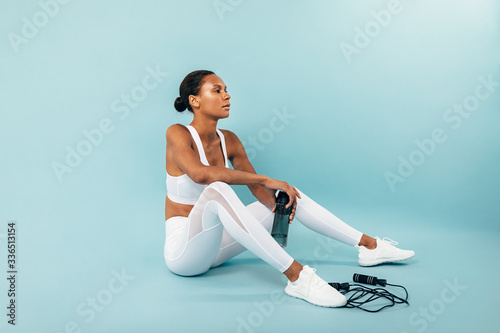 Tired woman in white sportswear sitting on a blue background. Female athlete holding water bottle in studio. photo