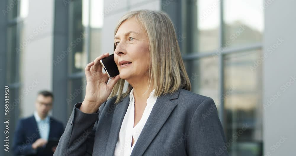 Close up of cheerful Caucasian beautiful businesswoman with gray hair talking on smartphone outdoors. Attractive happy woman speaking on mobile phone while chatting at street. Female using cellphone. Close up of cheerful Caucasian beautiful businesswoman with gray hair talking on smartphone outdoors. Attractive happy woman speaking on mobile phone while chatting at street. Female using cellphone.