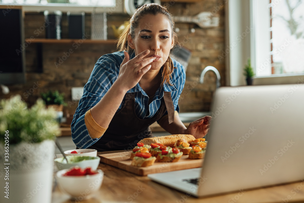 Young happy woman using laptop while preparing bruschetta in the kitchen. - obrazy, fototapety, plakaty 