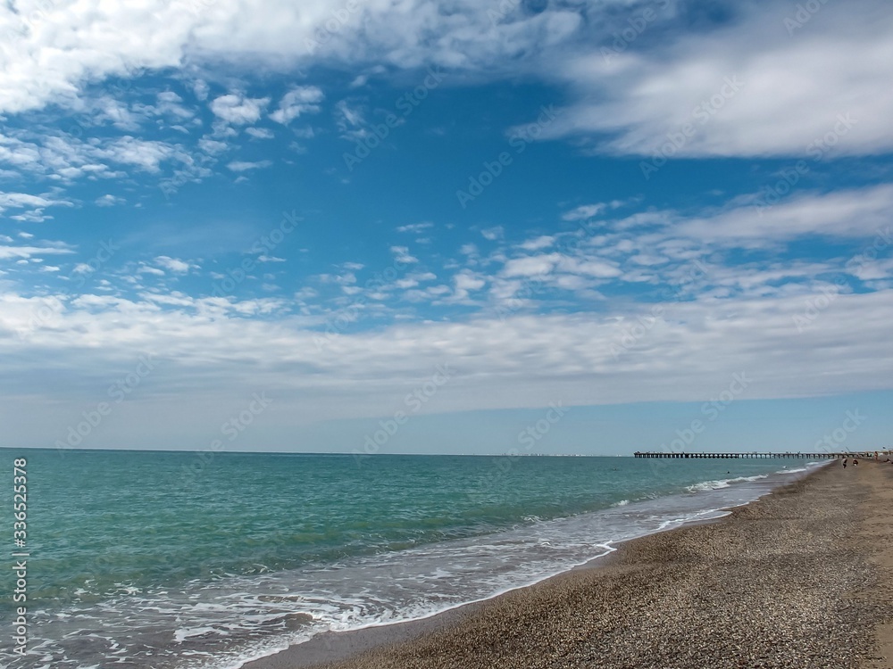 Blue sky with clouds over the green water of the sea and the coast of sand and pebbles.