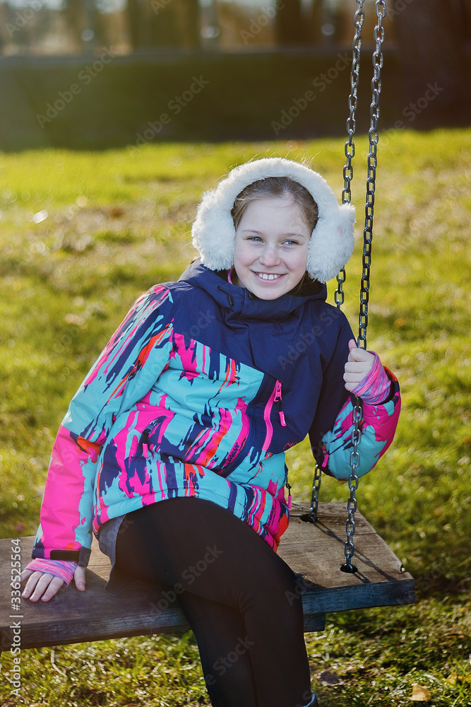 Child girl with blue eyes, white headphones on her head, in a spotted travel jacket, riding on a swing, holding on to an iron chain Child girl with blue eyes, white headphones on her head, in a spotted travel jacket, riding on a swing, holding on to an iron chain