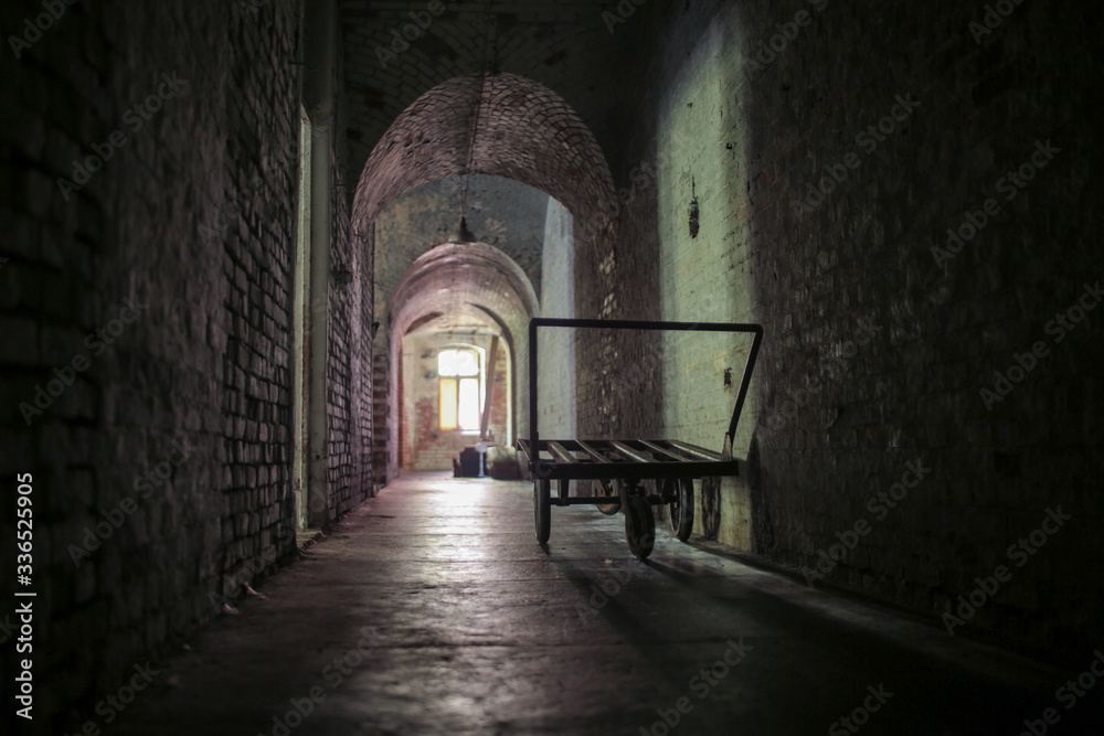 A long, dark corridor in an old abandoned fort. Stone walls. Shadow and light from small windows. 
There is an old, iron gurney against the wall