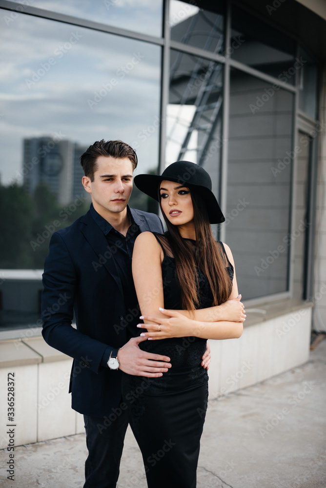 A beautiful, stylish pair of young people in black clothes and glasses stand against the background of an office building in the sunset. Fashion and style