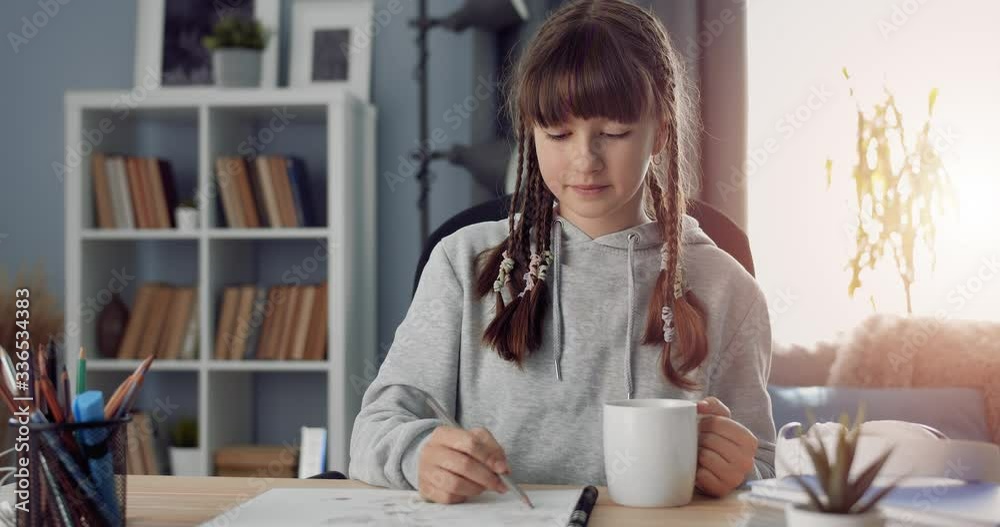 Cute female teenager in grey hoodie sitting at table with cup of tea and engaged in creativity. Happy girl with trendy hairstyle smiling and looking at camera. Cute female teenager in grey hoodie sitting at table with cup of tea and engaged in creativity. Happy girl with trendy hairstyle smiling and looking at camera.