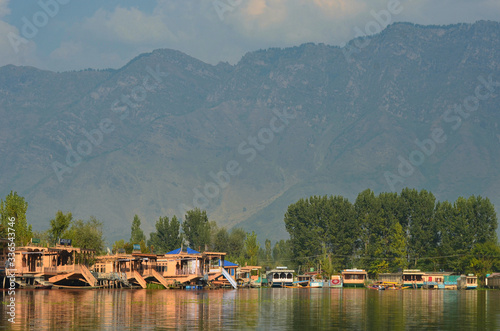 Wooden houseboats moored on Dal Lake in Kashmir. Their colours are reflected in the water. Trees line the shore, and mountains rise behind them to a cloudy sky.