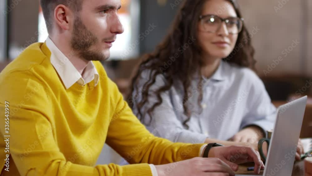 Tilt up shot of young caucasian businessman working on laptop and speaking with colleague while sitting with young woman at cafe table during coffee break Tilt up shot of young caucasian businessman working on laptop and speaking with colleague while sitting with young woman at cafe table during coffee break