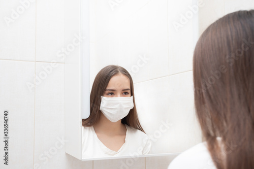 A girl in a medical mask looks in the mirror at home in the bathroom. Prevention and protection against diseases