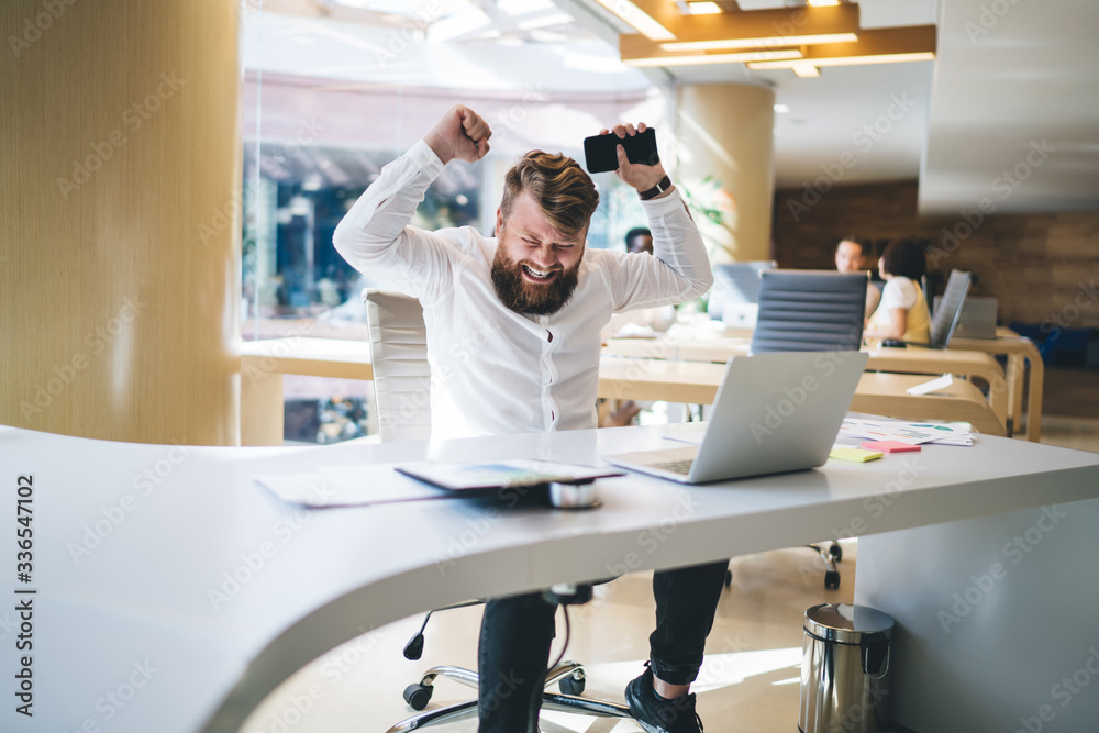 Obraz premium Accomplished male raising hands while celebrating completed startup project happy with received earning and new opportunity, satisfied businessman with cellphone gadget rejoicing at table desktop