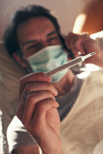 Coronavirus quarantine: young man lying in his bed with medical mask and thermometer