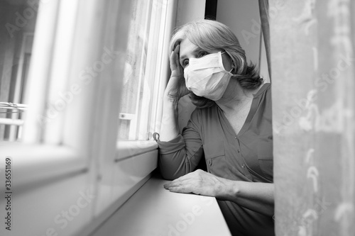 Sad senior woman wearing surgical mask looking through the window, covid19 pandemic  photo