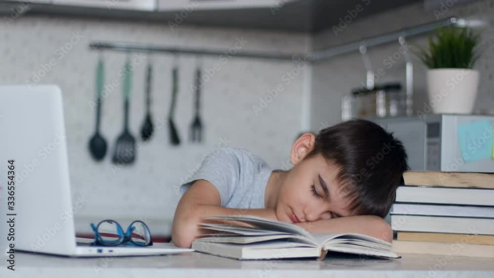 Tired schoolboy fell asleep doing homework sitting at a desk next to a laptop and textbooks Tired schoolboy fell asleep doing homework sitting at a desk next to a laptop and textbooks