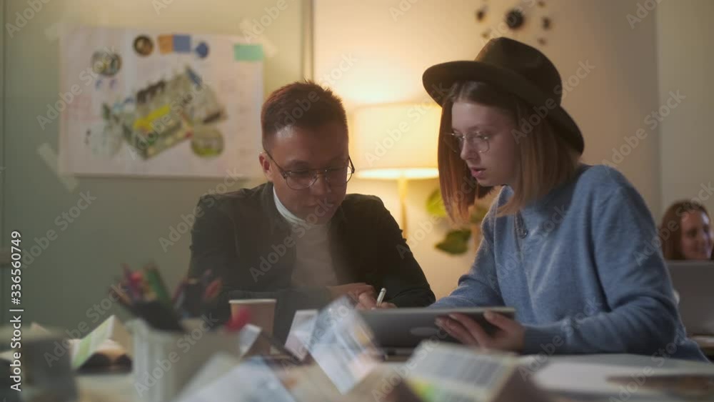 Two Stylish Diverse People Are Sitting At The Table While Energetically Discussing the Project. Evening Time. Drawing With Pen on Digital Tablet.