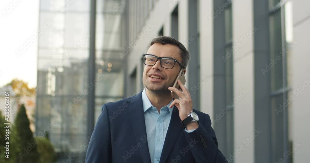 Cheerful Caucasian grey-haired middle-aged man in glasses talking on smartphone while walking the street. Handsome businessman rushing outdoors and speaking on mobile phone. Male speaking cellphone. Cheerful Caucasian grey-haired middle-aged man in glasses talking on smartphone while walking the street. Handsome businessman rushing outdoors and speaking on mobile phone. Male speaking cellphone.