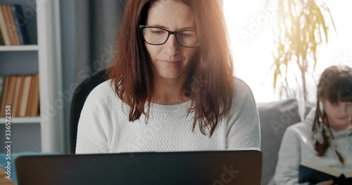 Mature woman in eyeglasses and white sweater using laptop for remote work while staying at home. Two children sitting quietly on couch and studying with book and laptop.