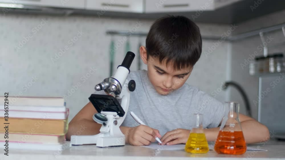 Close-up. A schoolboy teaches chemistry using a microscope and flasks with liquid and makes notes in a notebook. Homework. Close-up. A schoolboy teaches chemistry using a microscope and flasks with liquid and makes notes in a notebook. Homework.