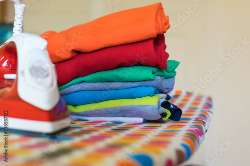A stack of clothes and a red and white iron on the Ironing board.