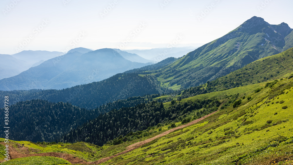 Fototapeta premium Green mountain slope. Layers of mountains in the haze during sunset. Krasnaya Polyana, Sochi, Caucasus, Russia.
