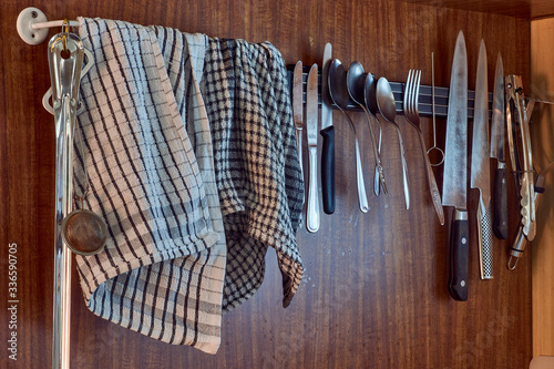 Knives and cutlery hang from a rail, beside tea towels which are draped on a rail; in a 70s kitchen with fake wood-grain brown vinyl walls.