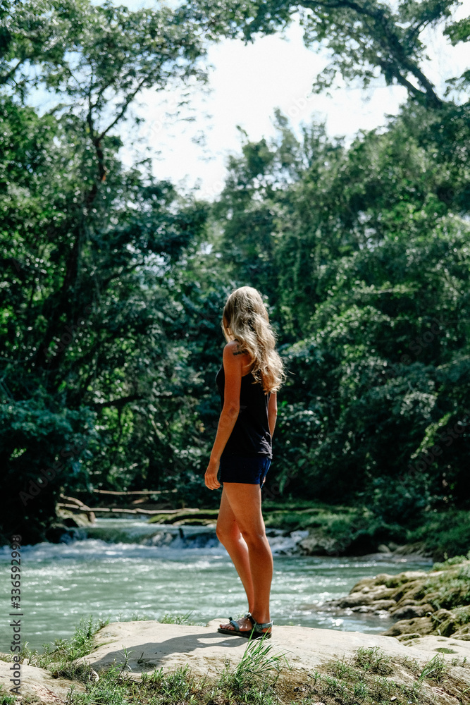 Tall blonde girl with long hair stands with her back and looks at the green forest in sunny weather. Travel