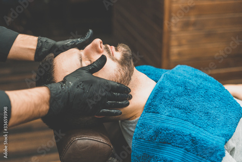 Barbershop. The client in the master’s chair in the barbershop, the barber applies oil and cosmetics to the client’s beard. Male beauty shop. Healthy lifestyle and beauty.
