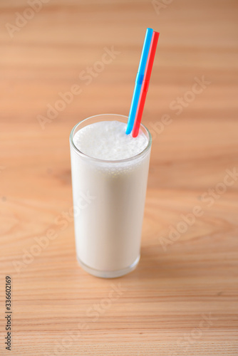 Vanilla milkshake in a tall galss with two plastic straws on light rustic wooden table.