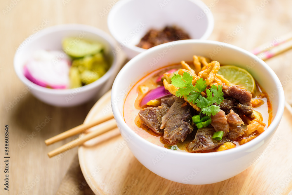 Northern Thai food (Khao Soi), spicy curry noodles soup with coconut milk and beef in a bowl on wooden background Northern Thai food (Khao Soi), spicy curry noodles soup with coconut milk and beef in a bowl on wooden background