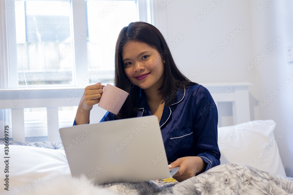 Young asian smiling woman working on laptop while holding a pink mug. Happy morning Young asian smiling woman working on laptop while holding a pink mug. Happy morning