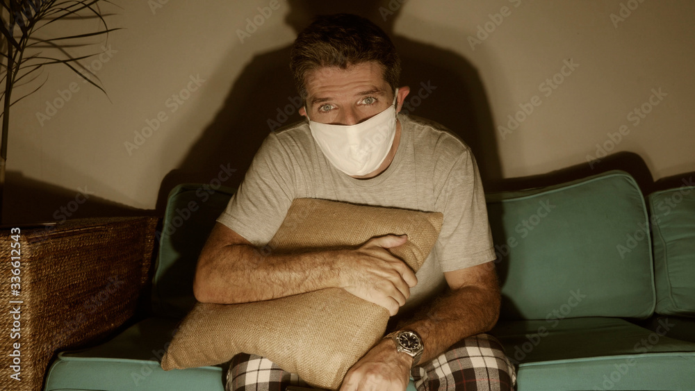 covid-19 virus home quarantine - young attractive scared and worried man in protective mask watching TV news about coronavirus pandemic crisis following stay home advise