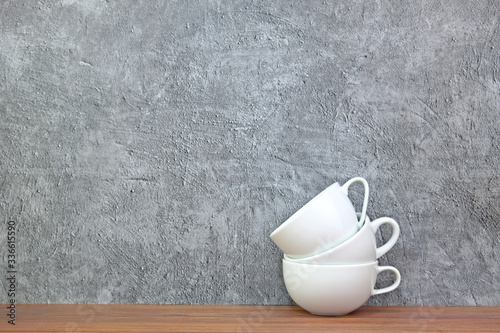 Three empty white coffee cups stacked on top of each other on wooden table with concrete background having copy space on the left.