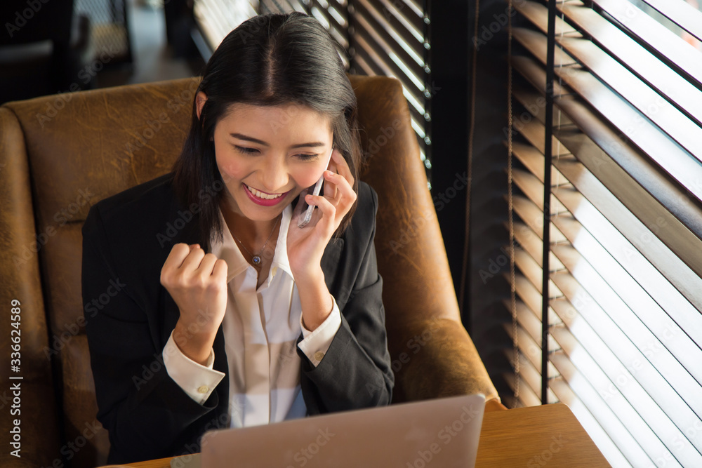 portrait charming young Asian woman in formal suit celebrating success with arms up while talking with cell phone in front of laptop on wooden table next the window portrait charming young Asian woman in formal suit celebrating success with arms up while talking with cell phone in front of laptop on wooden table next the window